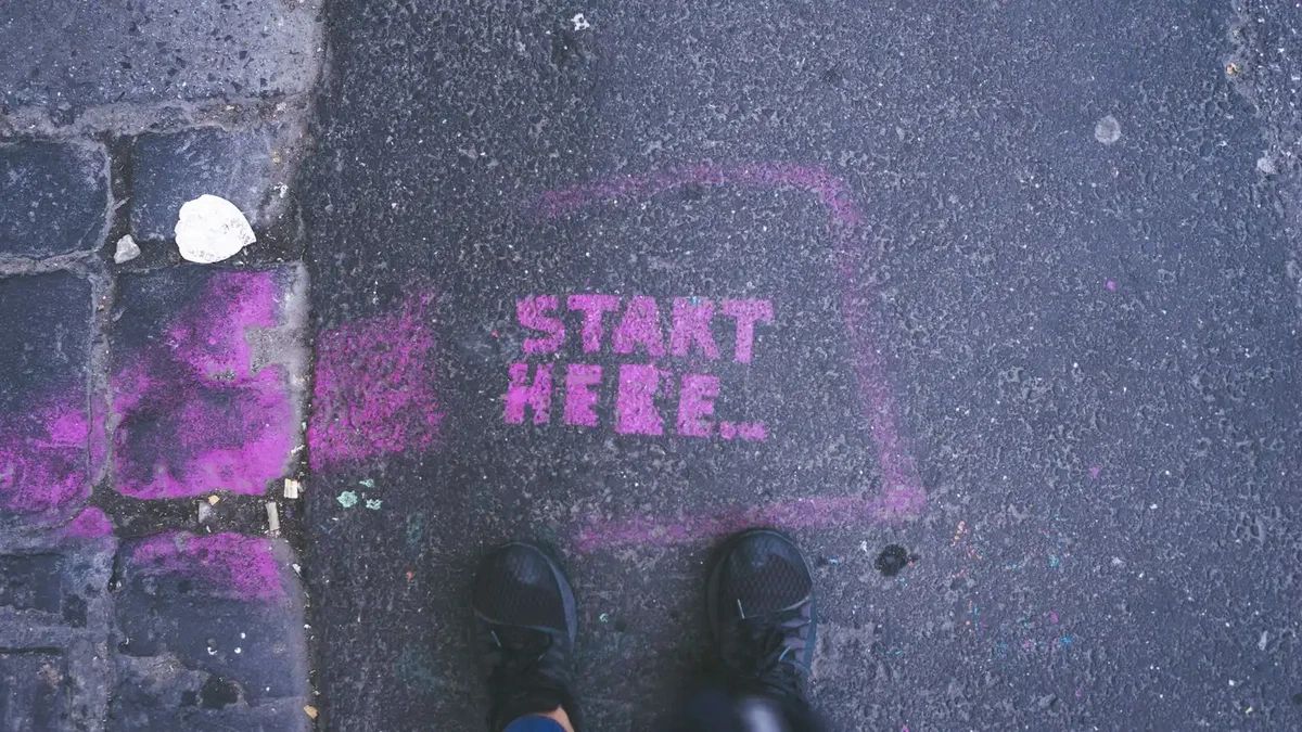 A pavement seen from above, with the words START HERE stencilled in bright pink spray paint inside a hand-drawn arrow, and a person's trainers standing just below the text.