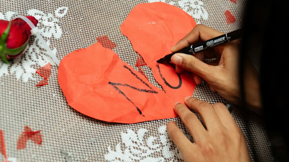 A hand writing the word NO in black marker across a torn red paper heart resting on white lace, a single red rose lying beside it.