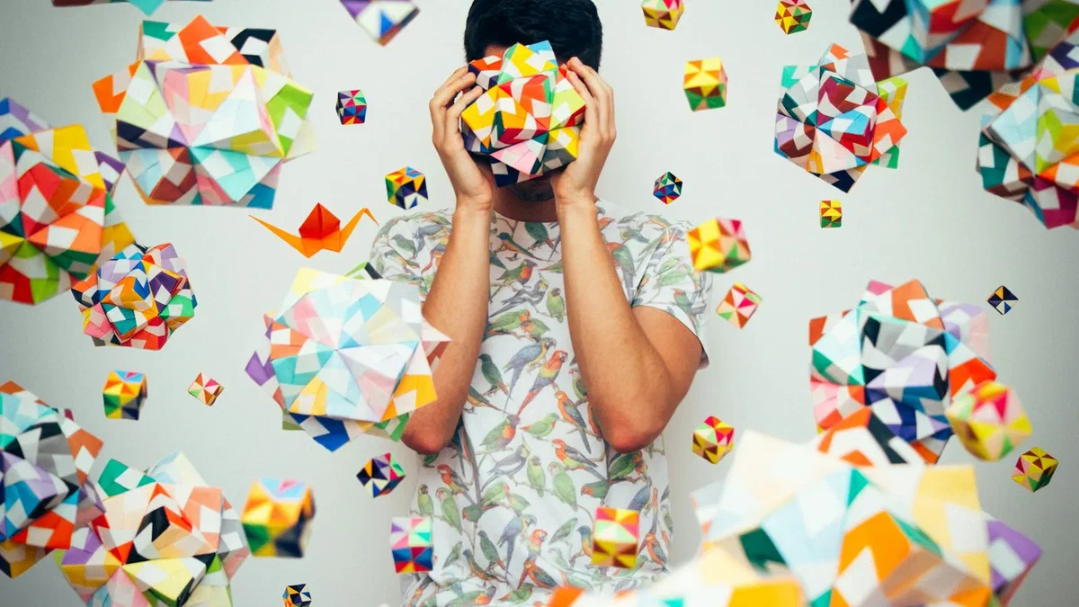 A man stands against a white wall holding a brightly coloured geometric origami ball over his face, surrounded by dozens of floating paper origami shapes in clashing colours.
