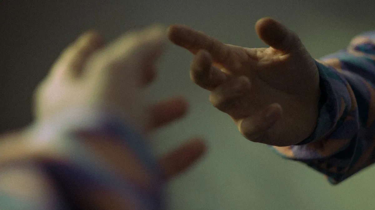 Two hands reaching towards each other in warm, low light, fingertips almost touching, against a soft green background.