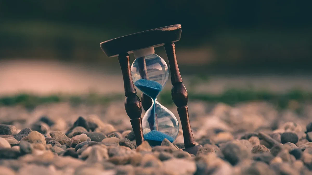 A small hourglass with blue sand standing upright on a bed of smooth grey and brown pebbles, the sand mid-flow from the top bulb to the bottom, against a softly blurred background.